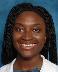 Andoh A woman with braided hair smiles at the camera. She is wearing a white coat over a patterned blue top, and the background is a solid blue color.