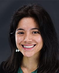 Becerra A young woman with long dark hair, wearing a green top and drop earrings, smiles at the camera against a dark, blurred background.