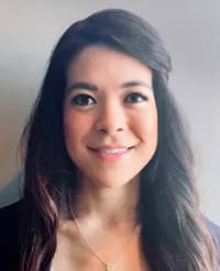 Chiou A woman with long dark hair, wearing a necklace and a dark top, smiles in front of a plain, light-colored background.