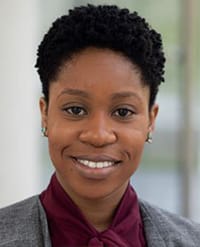 Ikpoh A woman with short curly hair smiles at the camera. She is wearing a gray blazer over a burgundy blouse and small earrings. The background is blurred and light-colored.