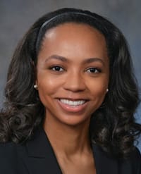 Livingston A woman with long, wavy dark hair, wearing a black blazer and a headband, smiles at the camera against a gray background.