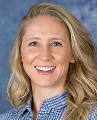 Naranjo A woman with long blonde hair smiles at the camera. She is wearing a blue and white checked shirt and stands in front of a blue, textured background.