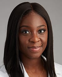Ocran A woman with long straight black hair and dark skin, wearing a white top and a gold necklace, smiles softly against a plain gray background.