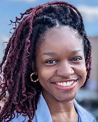 Oluwasola A young woman with long, burgundy-tinted braids smiles outdoors, wearing a light blue collared shirt and hoop earrings. The background is softly blurred with hints of sky and buildings.