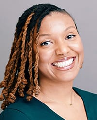 Propst A woman with medium brown skin and shoulder-length, twisted hair with lighter brown highlights is smiling at the camera. She is wearing a dark top and a delicate necklace, with a plain gray background behind her.