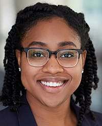 Simmons A smiling woman with dark braided hair, wearing glasses and a navy blazer, is pictured in a professional headshot with a blurred background.