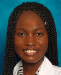 Sopeyin A woman with dark skin and long, twisted braids smiles at the camera. She is wearing a white coat and a striped shirt, with a blue background behind her.