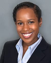 Thermozier A woman with dark braided hair, wearing a dark blazer and a light blue collared shirt, smiles in front of a plain gray background.
