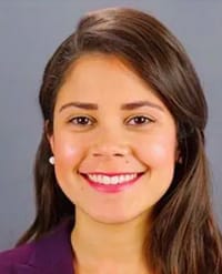 Villegas A woman with long brown hair, wearing a dark blazer and white stud earrings, smiles at the camera against a plain gray background.