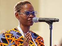 A woman with short hair and glasses speaks into a microphone. She is wearing a colorful patterned top and a white lanyard, standing indoors against a plain background.