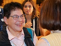 An older woman with short dark hair and glasses smiles warmly at another person, while a younger woman stands in the background, watching and smiling.