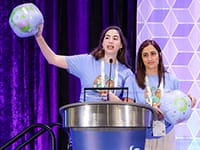 Two women in blue shirts stand at a podium on stage. One holds up an inflatable globe while speaking, and the other stands beside her. A geometric pattern and purple curtain are in the background.