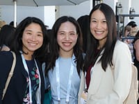 Three women smiling and standing close together at an outdoor event, wearing business attire and conference lanyards. There are other people and white umbrellas visible in the background.