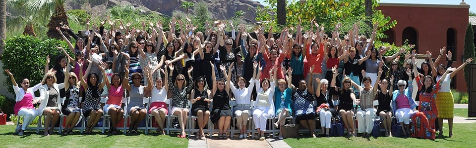 A large group of people, mostly women, are sitting and standing on benches outdoors, smiling and raising their arms in celebration, surrounded by greenery and palm trees.