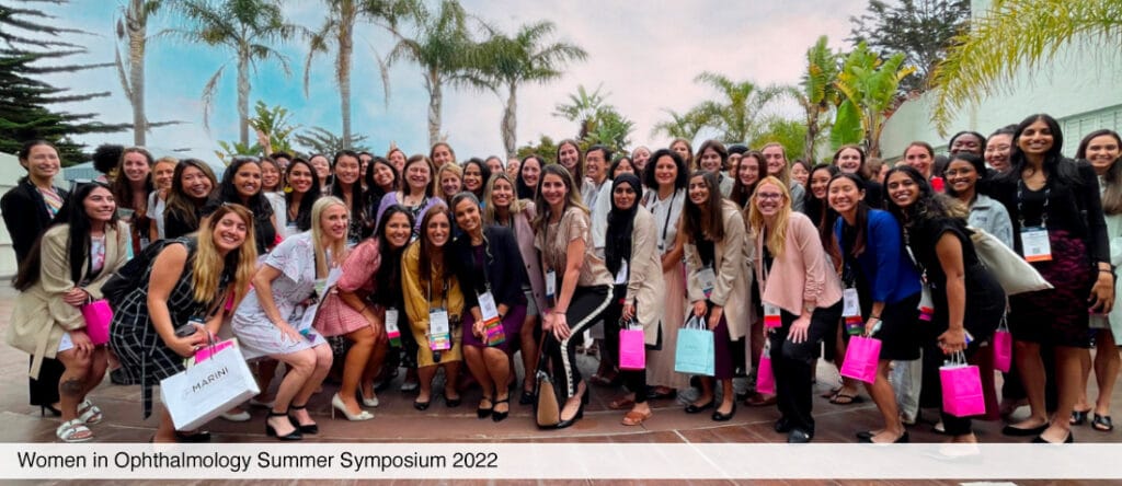 A large group of women stand smiling outdoors, many holding gift bags, at the Women in Ophthalmology Summer Symposium 2022, with palm trees and a cloudy sky in the background.