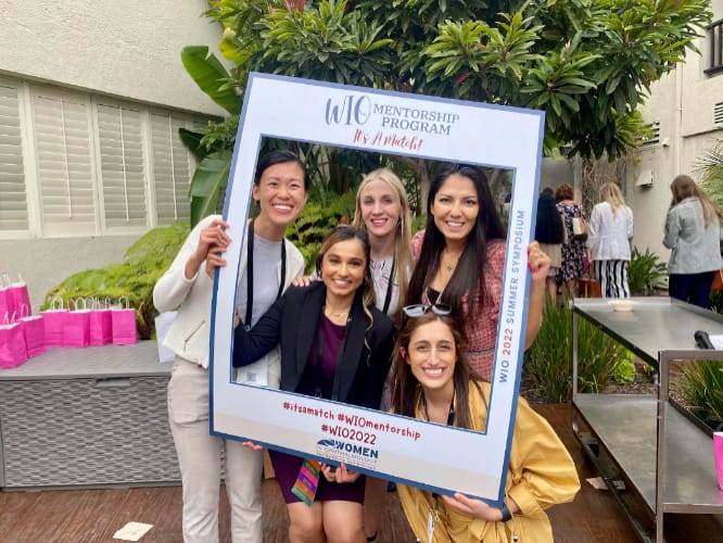 Five women smile and pose together outdoors, holding a large decorative frame promoting the WIO Mentorship Program. Other people and pink gift bags are seen in the background near greenery and a building.