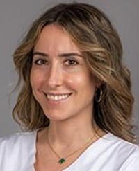 orešković-tall A woman with wavy, shoulder-length light brown hair is smiling. She is wearing a white top, gold hoop earrings, and a gold necklace with a small green pendant. The background is plain and gray.