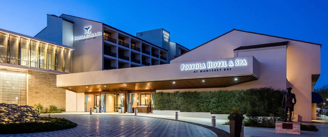 The Portola Hotel & Spa at Monterey Bay exterior at dusk, with modern architecture, illuminated signs, and a covered entrance surrounded by landscaping and a paved driveway.