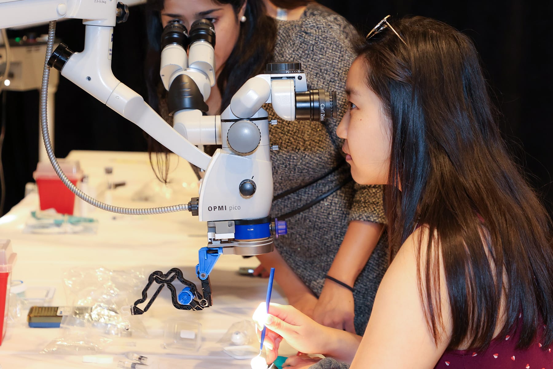 Two women use a microscope on a table with lab equipment. One woman looks through the eyepiece, while the other stands behind her, observing. The table holds various scientific tools and materials.