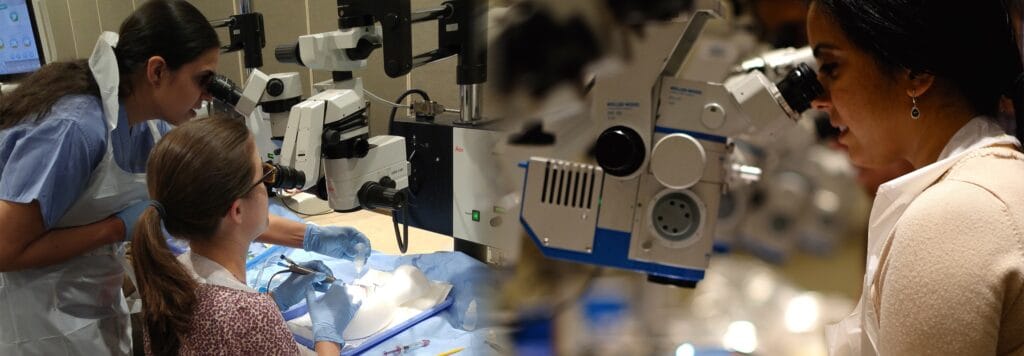 Three people, wearing gloves and aprons, use microscopes to observe samples in a laboratory setting. Various scientific equipment and tools are visible on the table.