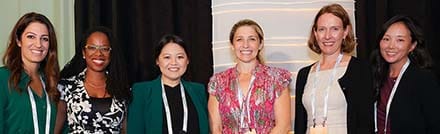 Six women stand side by side, smiling at the camera, dressed in business or professional attire with conference lanyards around their necks, in front of a dark curtain backdrop.