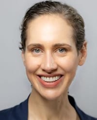 pershing A woman with light skin, blue eyes, and brown hair pulled back, smiling at the camera. She is wearing a dark-colored top and is set against a plain light gray background.