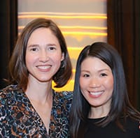 Two women smiling and posing together indoors, with a softly lit background featuring warm, horizontal lights.
