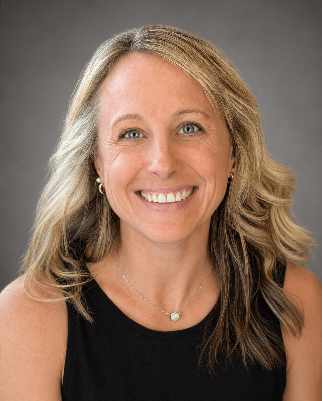 Slindquist Headshot 8.1.26 A smiling woman with wavy blonde hair wearing a black sleeveless top and a silver necklace, posed against a gray background.