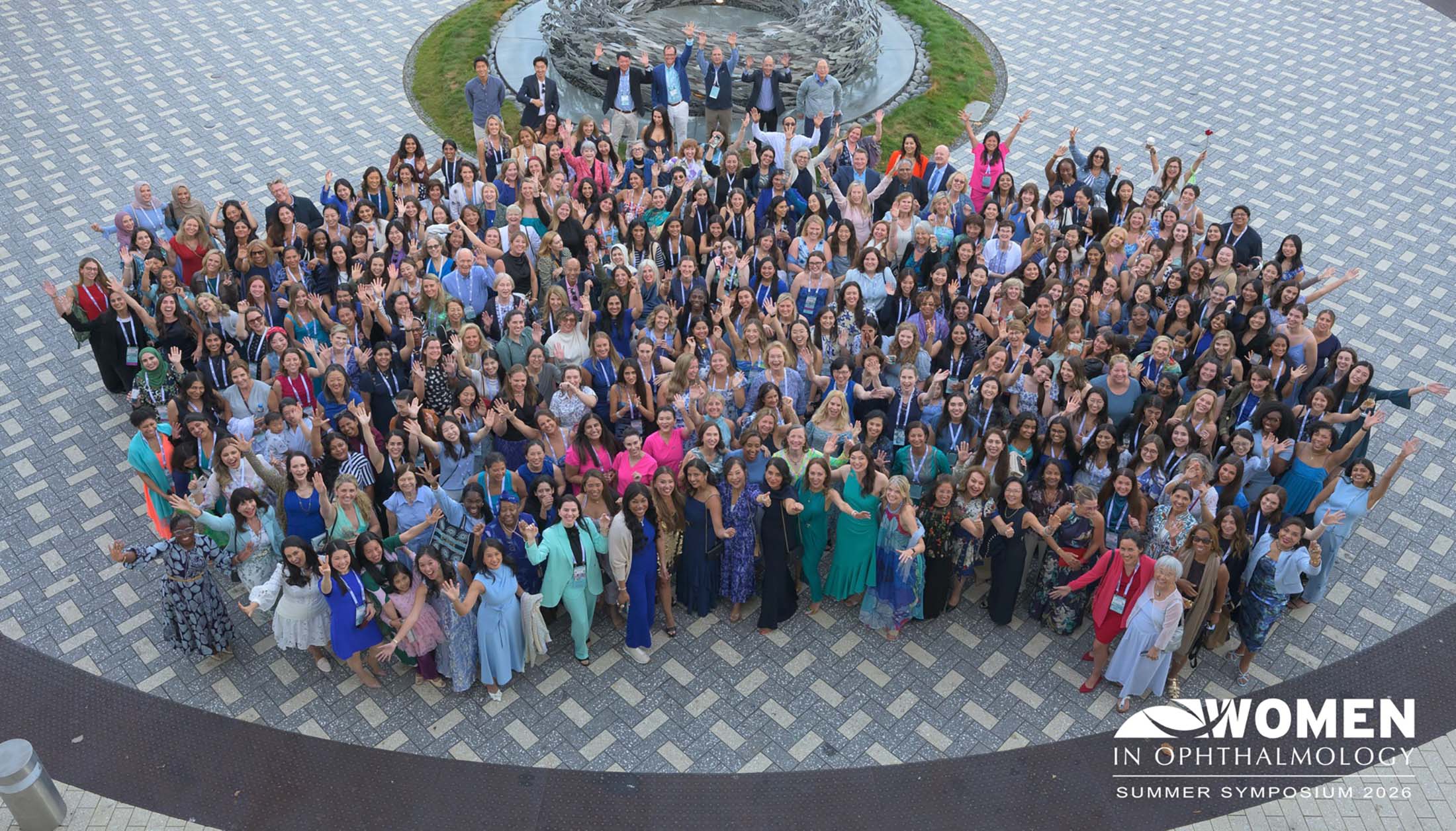 Large group of women and a few men stand outside, smiling and waving at the camera. They are attending the Women in Ophthalmology Summer Symposium 2023. A decorative fountain and patterned stone walkway are visible.
