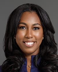 desilva Portrait of a woman with long, wavy dark hair, wearing a navy blue top and a small gold necklace, smiling at the camera with a neutral grey background.