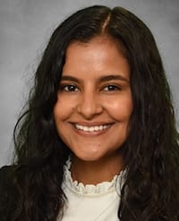 khanal A woman with long, wavy dark hair smiles at the camera. She is wearing a white top with a ruffled collar and a dark blazer. The background is a simple gray gradient.