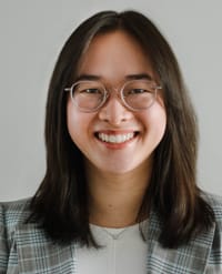 Anne Xuan-Lan Nguyen, MD Smiling person with long dark hair, wearing round glasses, a light plaid blazer, and a white top, standing in front of a plain light background.