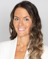 Alexander Headshot of a woman with long, wavy brown hair smiling at the camera, wearing a white blazer and a delicate necklace.